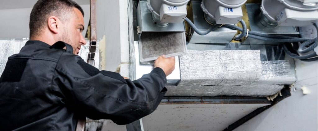 A professional furnace tech is opening the furnace and taking out the blower to clean the inner workings of the furnace. This is a forced air furnace that is commonly found in homes in Alberta. The furnace tech is wearing blue coveralls.